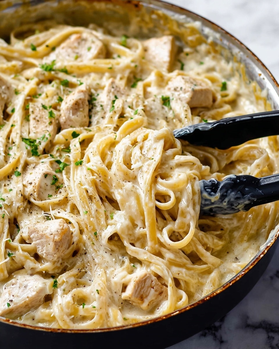 The image shows a close-up of a creamy chicken alfredo pasta in a pan. The dish has long, flat pasta noodles coated in a rich, pale cream sauce with smooth texture. There are several pieces of white chicken chunks mixed evenly throughout the noodles. The dish is sprinkled with small bits of green parsley and black pepper for color contrast. A pair of black tongs holds the pasta gently from the bottom right corner. The pan has a light scuff mark at the edges, and the background features a white marbled texture. photo taken with an iphone --ar 4:5 --v 7