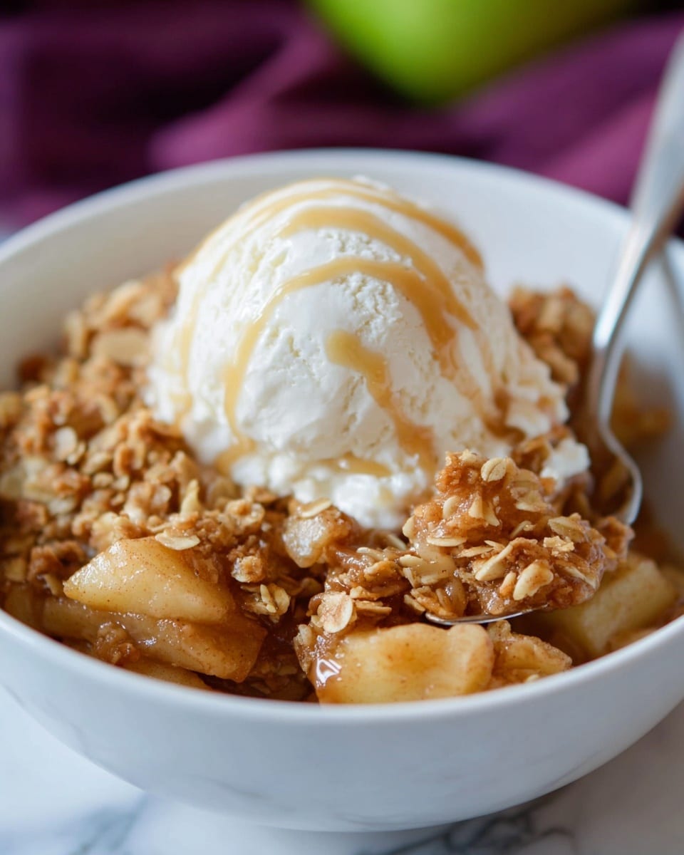 The image shows a white bowl filled with a warm apple crisp dessert. The bottom layer has chunky, soft cooked apple pieces coated in a golden-brown cinnamon sauce, giving a shiny texture. On top of the apples is a thick, crunchy oat crumble that looks golden with a rough texture. Above this is a large scoop of smooth, white vanilla ice cream that is slightly melting and drizzled with light caramel sauce. A silver spoon is partially inserted into the bowl, touching the apple crisp near the front. The bowl is placed on a white marbled surface with some out-of-focus colors in the background. photo taken with an iphone --ar 4:5 --v 7