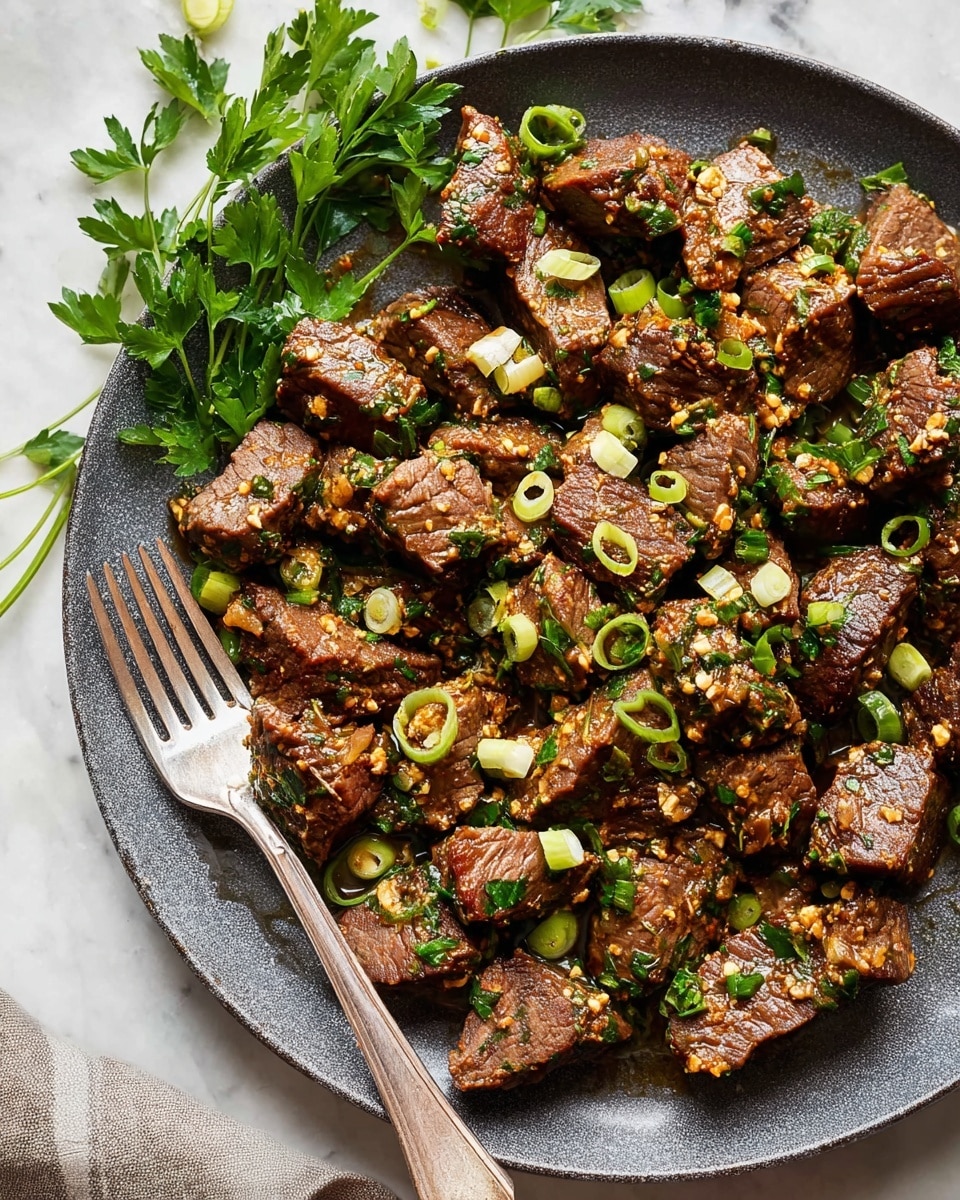 A dark gray round plate holds many pieces of cooked beef cubes scattered evenly. Each beef cube is covered with small bits of garlic and chopped green herbs, giving a textured look. Thin slices of fresh light green spring onions are mixed on top and around the beef pieces, adding soft green circles and rings. A silver fork rests at the left side of the plate on a white marbled surface, with a few sprigs of fresh flat green parsley leaves spread above it for decoration. The beef shines slightly with a glossy light brown sauce coating. Photo taken with an iphone --ar 4:5 --v 7