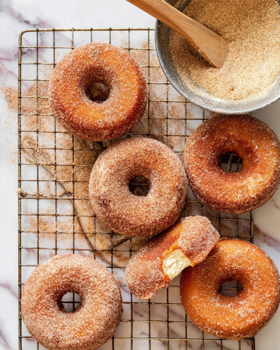 A top view of seven cinnamon sugar-coated donuts arranged on a wire cooling rack, each donut with a golden-brown color and a rough sugar texture on top, one donut showing a single bite taken from the edge revealing its soft inside, accompanied by a round metal bowl filled with cinnamon sugar mixture and a wooden spatula resting on it, all set on a white marbled surface photo taken with an iphone --ar 4:5 --v 7
