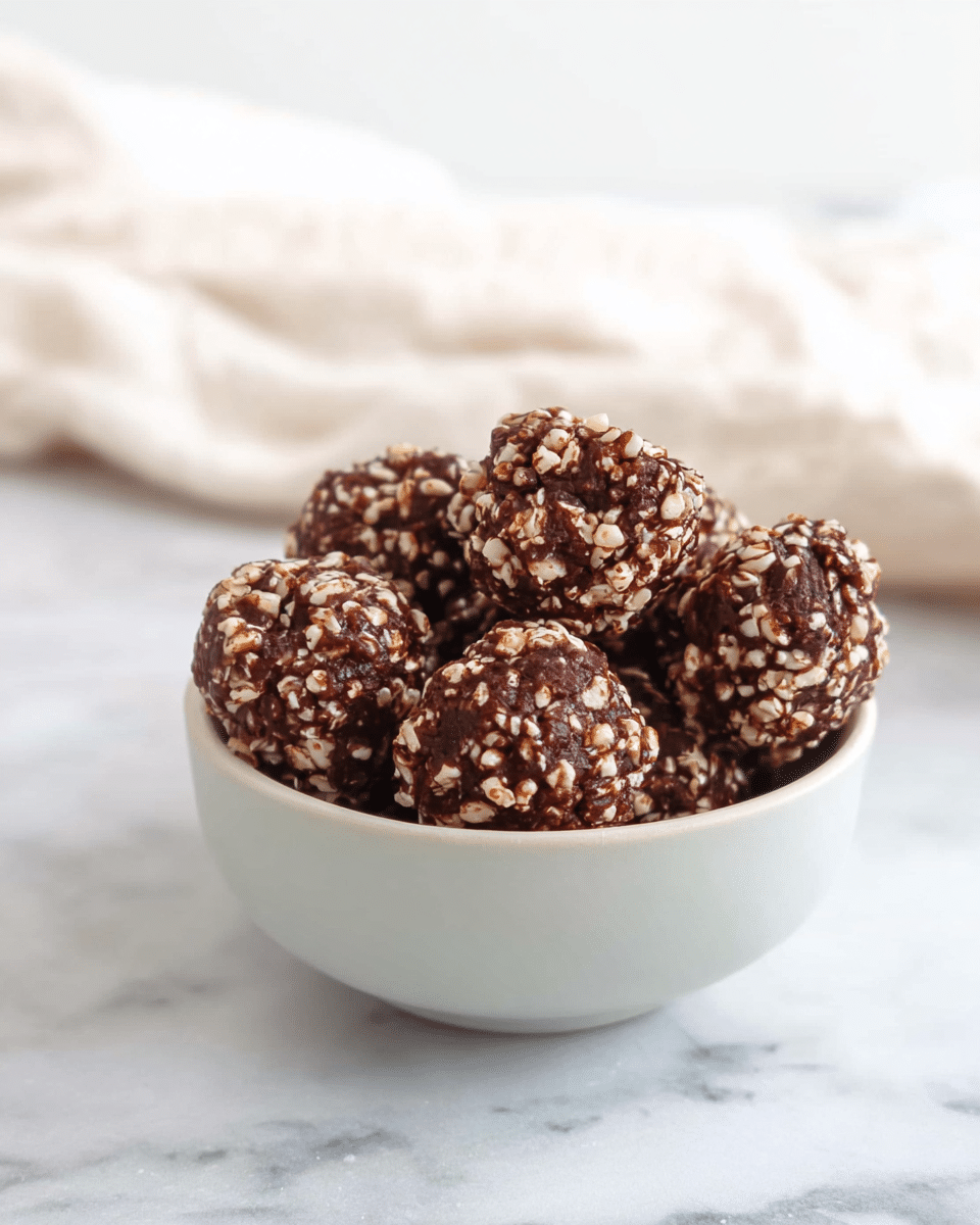 A small white bowl filled with round, bite-sized chocolate balls covered in small, light-colored puffed rice pieces, giving them a bumpy texture. The chocolate has a rich dark brown color, and the puffed rice bits are scattered evenly all over each ball. The bowl sits on a white marbled surface with a soft, blurred white cloth in the background. photo taken with an iphone --ar 4:5 --v 7