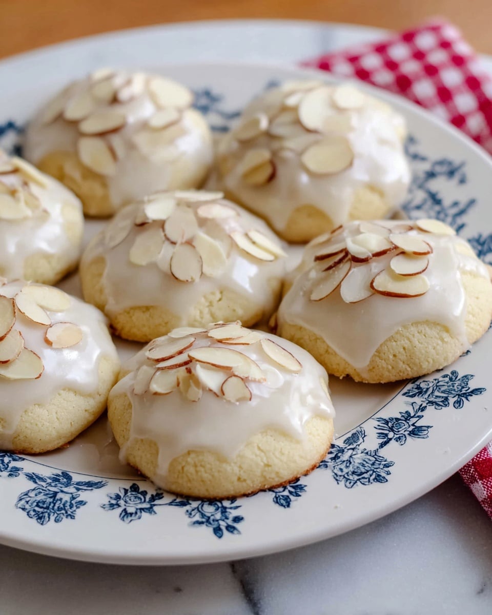 The image shows seven soft round cookies arranged on a white plate with a simple blue floral pattern. Each cookie has one layer made of pale dough that is thick and slightly puffy. The top of each cookie is covered with a smooth, white glaze that drips slightly down the sides. On top of the glaze, there are thinly sliced almond pieces, scattered evenly across each cookie. The plate is placed on a white marbled surface with a red and white checkered cloth visible in the background on the right side. photo taken with an iphone --ar 4:5 --v 7