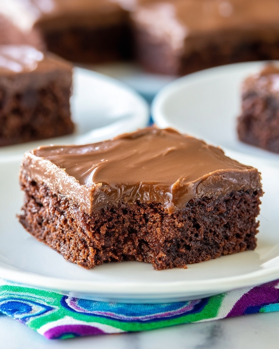 A close-up view of a single square piece of chocolate brownie with a bite taken out of its front side, showing a moist, dense, and slightly crumbly dark brown interior. The brownie has one thick layer of smooth, glossy, milk chocolate frosting spread evenly on top, with a shiny surface and subtle textured edges. The brownie rests on a round white plate positioned on a colorful patterned cloth with blue, green, purple, and red circular shapes. The background includes soft-focused pieces of similarly frosted brownies on white plates, all set against a white marbled surface. Photo taken with an iphone --ar 4:5 --v 7