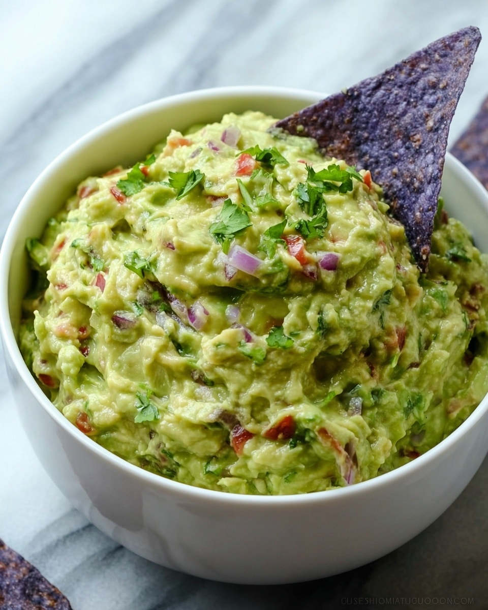 A close-up view of a white bowl filled with creamy guacamole showing a rough texture with visible small chunks of avocado, red onion, green herbs, and tiny pieces of tomato, all mixed together creating a greenish-yellow base with colorful specks. The bowl is placed on a white marbled surface, and the background is softly blurred with warm brown tones, giving focus to the bowl. Photo taken with an iphone --ar 4:5 --v 7