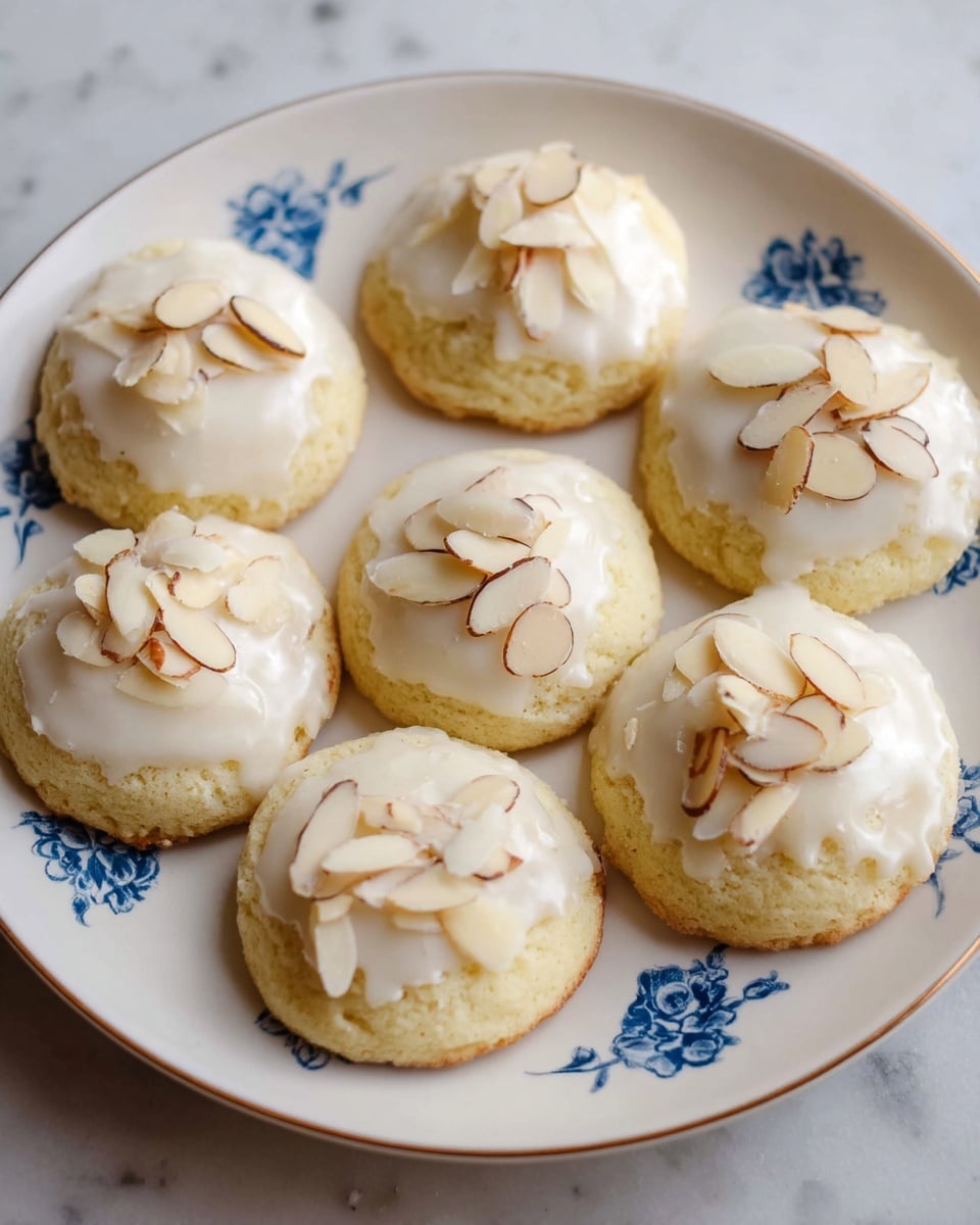 Seven round cookies are placed on a white plate with a simple blue floral pattern, all set on a white marbled surface. Each cookie has two layers: a soft, light yellow base with a slightly rough texture, topped with a smooth white icing layer that covers the top evenly. On top of the icing, thin almond slices with light brown edges are scattered, adding a slight crunch and contrast in color. The cookies are arranged close together in a casual circular form. photo taken with an iphone --ar 4:5 --v 7