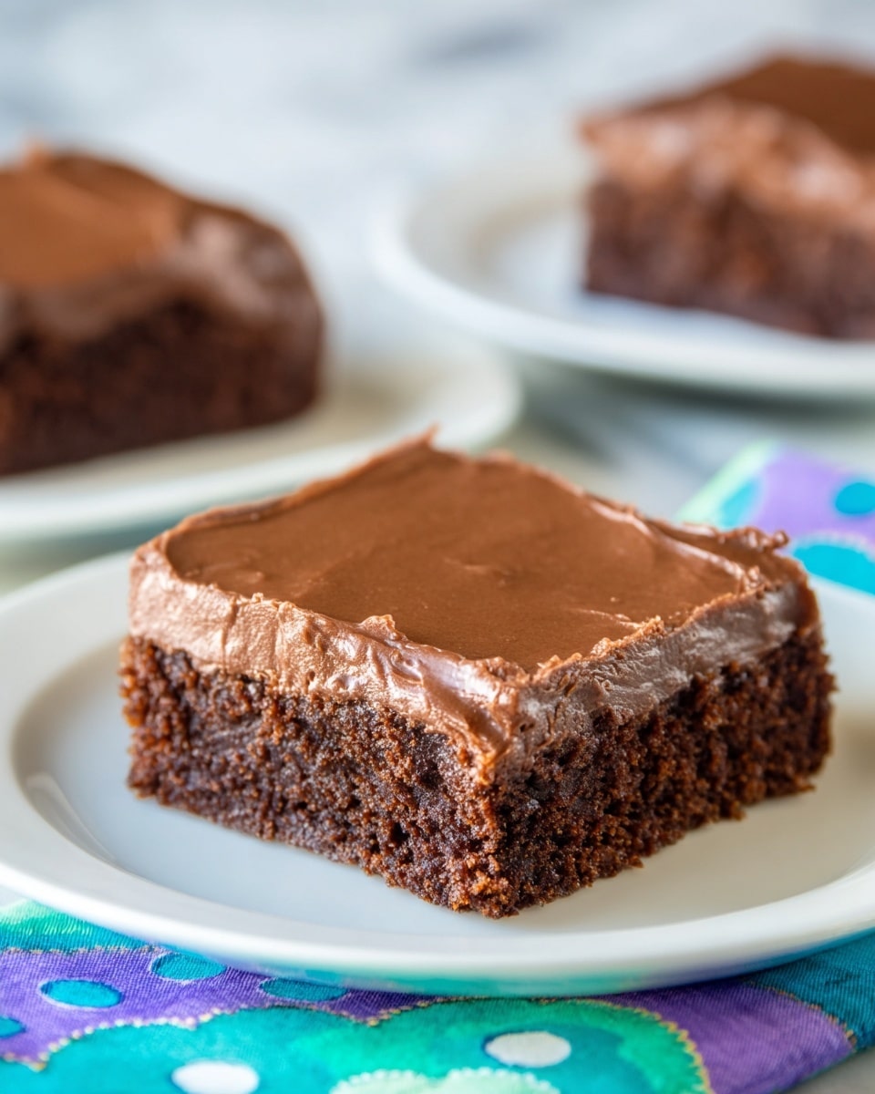 The image shows a close-up of a single square piece of chocolate brownie on a white plate. The brownie has two layers: the bottom layer is dense and dark brown with a slightly crumbly texture, while the top layer is a thick, smooth, and glossy chocolate frosting that covers the entire surface of the brownie evenly. The plate sits on a colorful cloth with blue, green, and purple circular patterns, all set against a white marbled background. In the blurred background, there are two more pieces of brownies on white plates. photo taken with an iphone --ar 4:5 --v 7