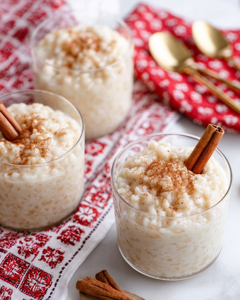 The image shows three clear glass cups filled with creamy rice pudding. Each cup has small textured grains of rice mixed in a thick, white custard-like base sprinkled lightly with brown cinnamon powder. A single brown cinnamon stick is placed inside each cup, sticking out from the creamy top layer. The cups are arranged on a white marbled surface covered partly by a white cloth with red patterns. A stack of folded red and white cloth napkins is nearby, along with two gold spoons and a few loose cinnamon sticks visible in the background. Photo taken with an iphone --ar 4:5 --v 7