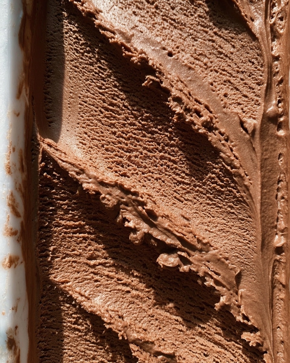 A white bowl holds four scoops of rich, dark brown chocolate ice cream, arranged closely together with some of the ice cream melting into a smooth pool at the bottom. The scoops have a soft, creamy texture with small ridges and melting edges. A shiny silver spoon rests in the bowl, touching the bottom scoop. The background shows a white marbled texture under the bowl. photo taken with an iphone --ar 4:5 --v 7