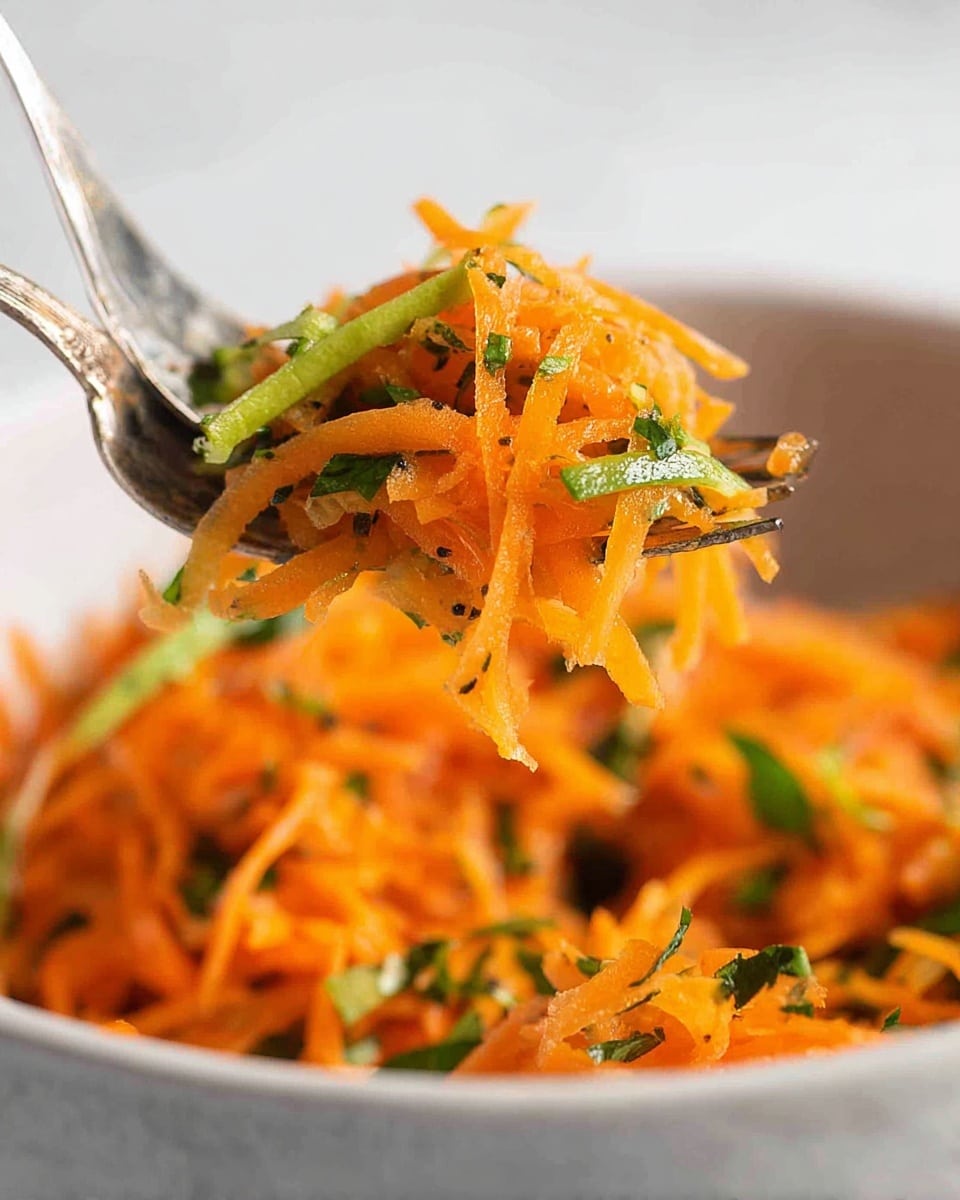 A dish in a light brown bowl shows a mix of thinly shredded orange carrots and sliced green herbs or scallions, evenly combined to create a bright, fresh look. The carrot strands form the main layer, with scattered green pieces adding contrast on top and throughout. Two silver spoons rest in the bowl, one leaning inside on the left side and the other set on the right, with the bowl placed on a soft gray cloth and a white marbled surface in the background. photo taken with an iphone --ar 4:5 --v 7