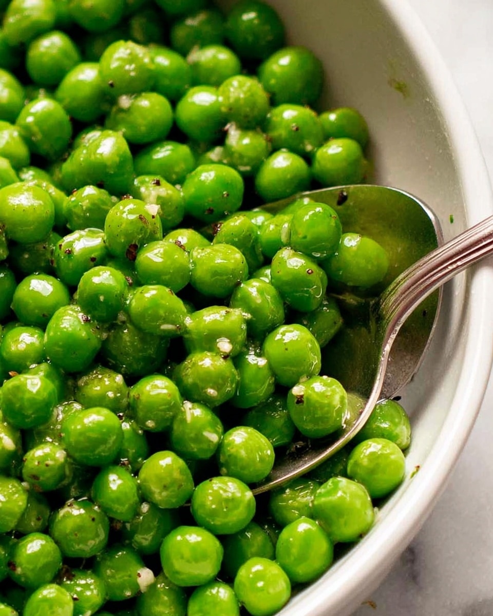 A close-up view of a white bowl filled with bright green peas that look fresh and juicy. The peas are glossy and slightly wet, with tiny specks of black pepper and small bits of garlic or seasoning scattered over them. A silver spoon is partially buried in the peas, showing some peas resting on the spoon's surface. The background is a white marbled texture, and the lighting highlights the shiny and vibrant color of the peas. photo taken with an iphone --ar 4:5 --v 7