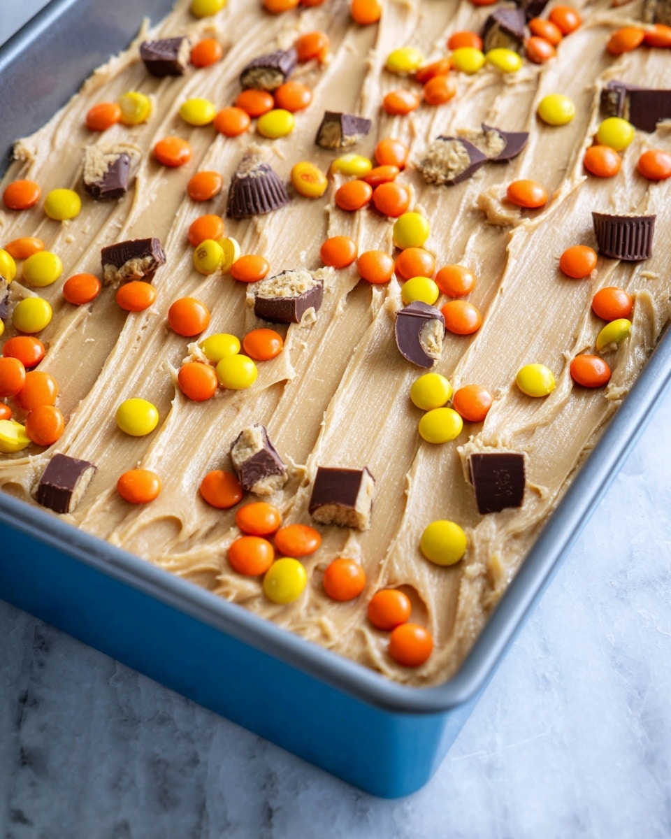 A close-up of a blue baking pan filled with a thick, smooth, light tan layer of cookie dough that has been lightly textured with curved lines across the surface. The top layer is sprinkled evenly with small, bright orange, yellow, and dark brown candy-coated chocolates, along with halved and whole mini chocolate peanut butter cups scattered throughout. The pan is set on a white marbled surface that softly contrasts with the colors of the cookie dough and candies. photo taken with an iphone --ar 4:5 --v 7
