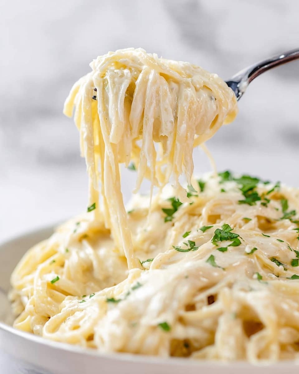 A close-up view of creamy white alfredo pasta with long noodles coated in a thick, smooth, and glossy white cheese sauce. Some green chopped herbs are sprinkled on top, adding small touches of color throughout. A fork is lifting a twisted bundle of the pasta above a white plate filled with more of the same creamy noodles against a blurred white marbled background. The texture looks rich and velvety with noodles slightly shiny from the sauce. photo taken with an iphone --ar 4:5 --v 7