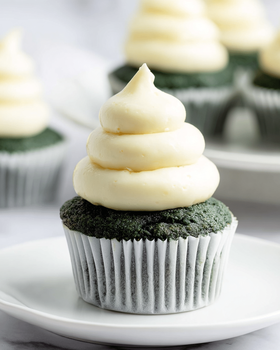A close-up of a single dark green cupcake in a white paper liner with a moist, slightly textured surface. On top is a tall swirl of smooth, pale yellow frosting with a soft, creamy texture, standing in three thick layers that taper to a point at the top. The cupcake rests on a plain white plate, all set on a white marbled background. In the blurred background are a few more green cupcakes in white liners, slightly out of focus. photo taken with an iphone --ar 4:5 --v 7