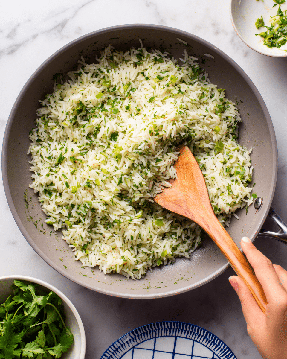 In a large gray pan, there is a single main layer of cooked rice mixed with finely chopped green herbs, making the rice look greenish with a slightly fluffy texture. A wooden spatula is scooping some of the rice from the center, held by a woman's hand visible at the bottom right. Around the pan, part of a white bowl with fresh green leafy herbs and a white plate with a blue grid pattern are seen on a white marbled surface. Photo taken with an iphone --ar 4:5 --v 7