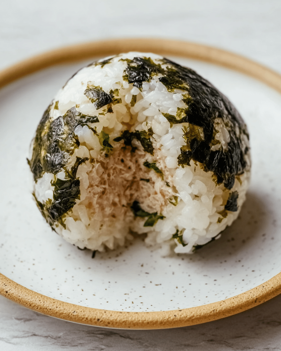 A close-up of a round rice ball with a soft and slightly sticky texture, covered in small pieces of dark green seaweed throughout its outside layer. The inside features a light brown, moist filling, which looks smooth and finely packed. The rice grains are white and distinct, surrounding the filling completely. This rice ball sits alone on a white plate with a speckled beige rim, placed on a white marbled texture surface. photo taken with an iphone --ar 4:5 --v 7