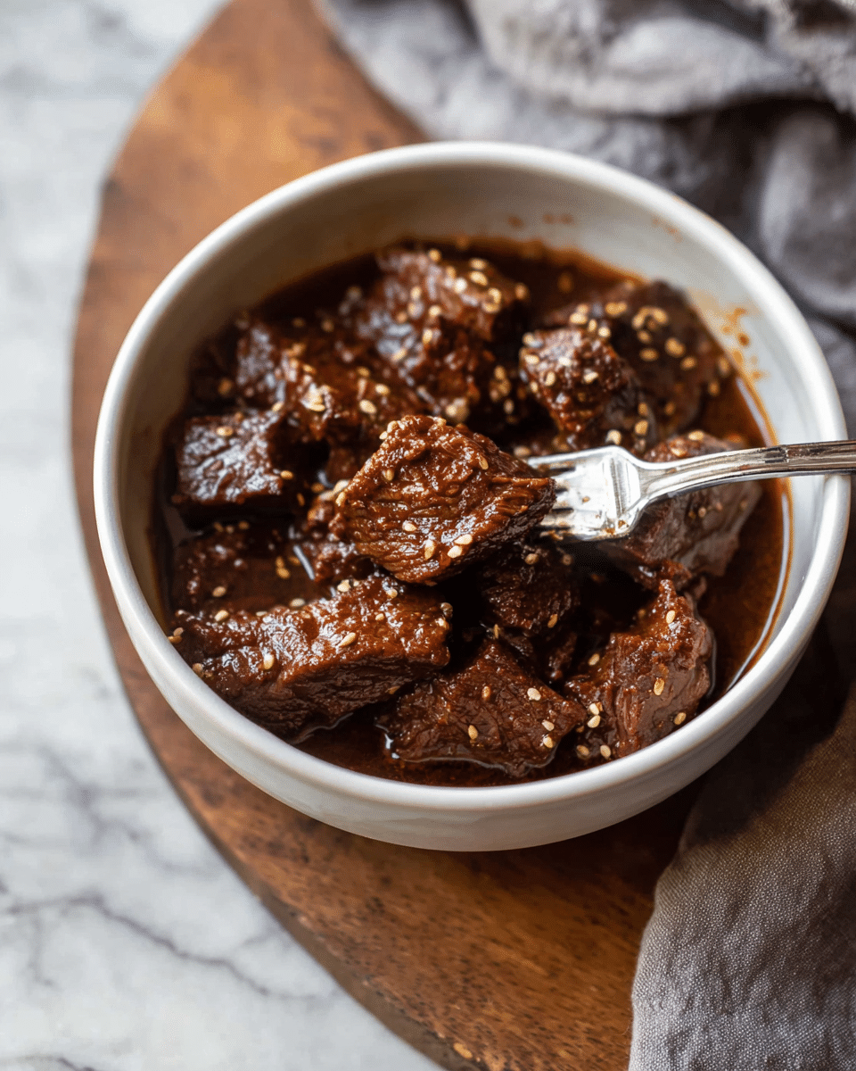 The dish is served in a white bowl filled with dark brown, tender chunks of meat, soaked in a rich, glossy sauce that has a slightly oily texture. The meat pieces are uneven in shape and coated in the sauce, with small sesame seeds sprinkled on top, adding a touch of light tan color and texture. A silver fork is partially inserted into the meat, lifting one piece from the bowl. The bowl rests on a white marbled surface with a hint of a soft gray cloth beneath it and a wooden board nearby. The photo taken with an iphone --ar 4:5 --v 7