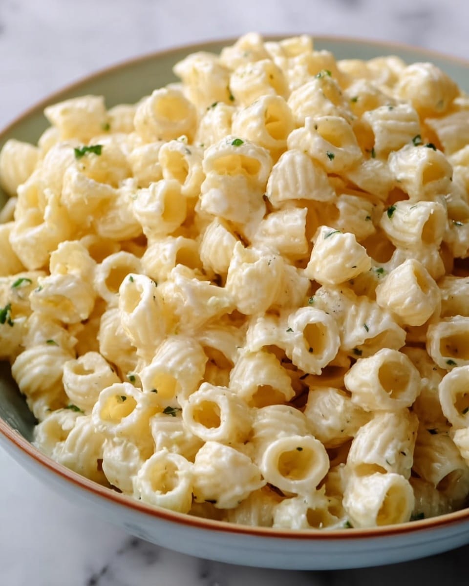 A close-up view of a white bowl with a blue rim filled with a creamy macaroni salad. The dish shows small elbow macaroni coated in a thick white mayonnaise-like sauce, mixed with slices of hard-boiled eggs, which add white and yellow layers throughout. There is a light sprinkling of red paprika on top, giving small red dots that contrast with the creamy white salad. The background has a white marbled texture with a slightly blurred look showing another bowl with more macaroni salad behind. photo taken with an iphone --ar 4:5 --v 7