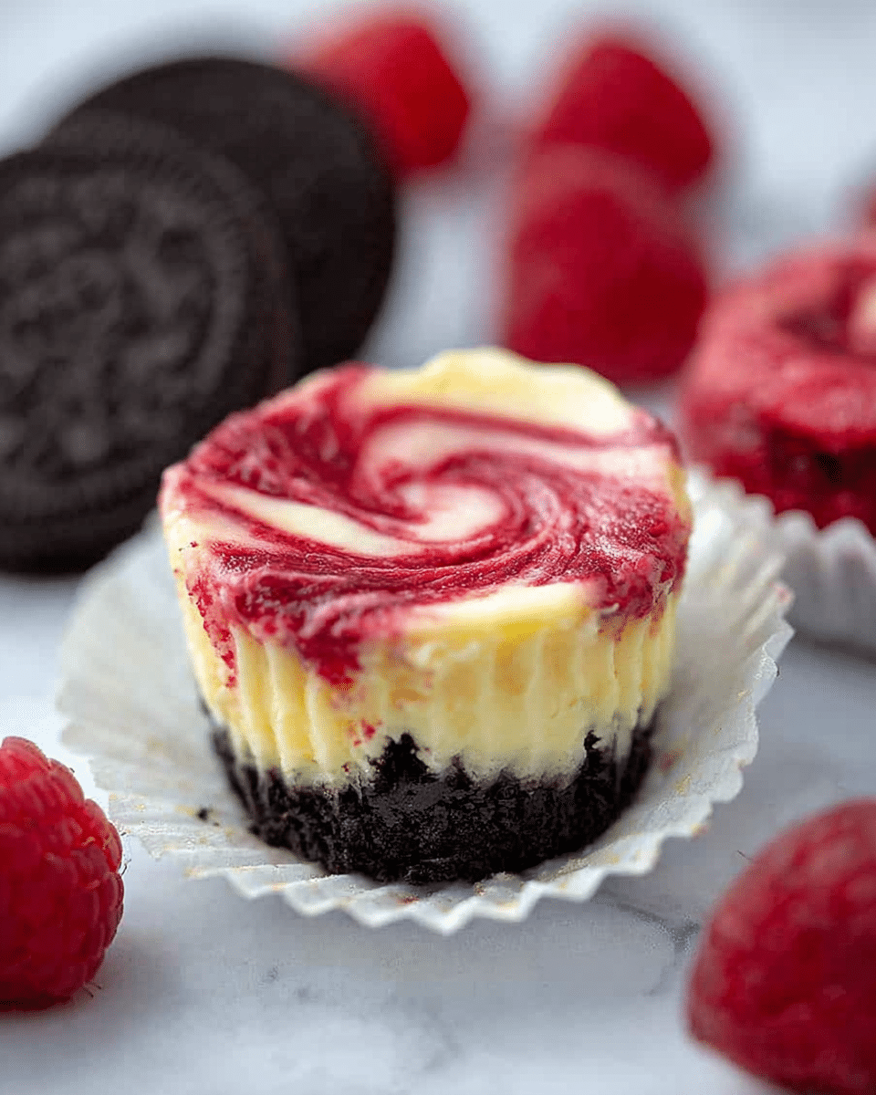 A small cheesecake cup with three visible layers sits on a white marbled surface. The bottom layer is a dark, crumbly Oreo crust, topped with a thick, creamy pale yellow cheesecake layer. The top layer is a smooth, swirled bright red raspberry sauce lightly mixed into the cheesecake, creating a marbled pattern. The cheesecake is in a partially peeled white paper liner, with two Oreo cookies and several red raspberries blurred in the background. Photo taken with an iphone --ar 4:5 --v 7