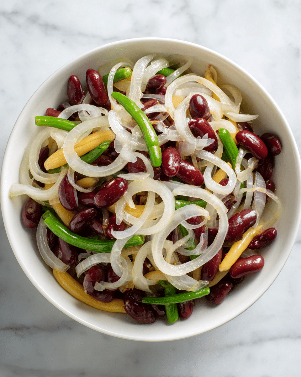 A white bowl holds a bright salad made of green beans, yellow wax beans, and small red kidney beans mixed together. On top, thin rings of white onion are spread evenly, adding contrast to the mixture. The salad looks fresh and slightly shiny, suggesting a light dressing. A wooden spoon rests inside the bowl, partly submerged in the salad. The bowl sits on a light textured beige cloth with a glass of amber liquid behind it and a piece of brown bread on a white plate with a blue checkered napkin on the side. The background is a white marbled texture. photo taken with an iphone --ar 4:5 --v 7