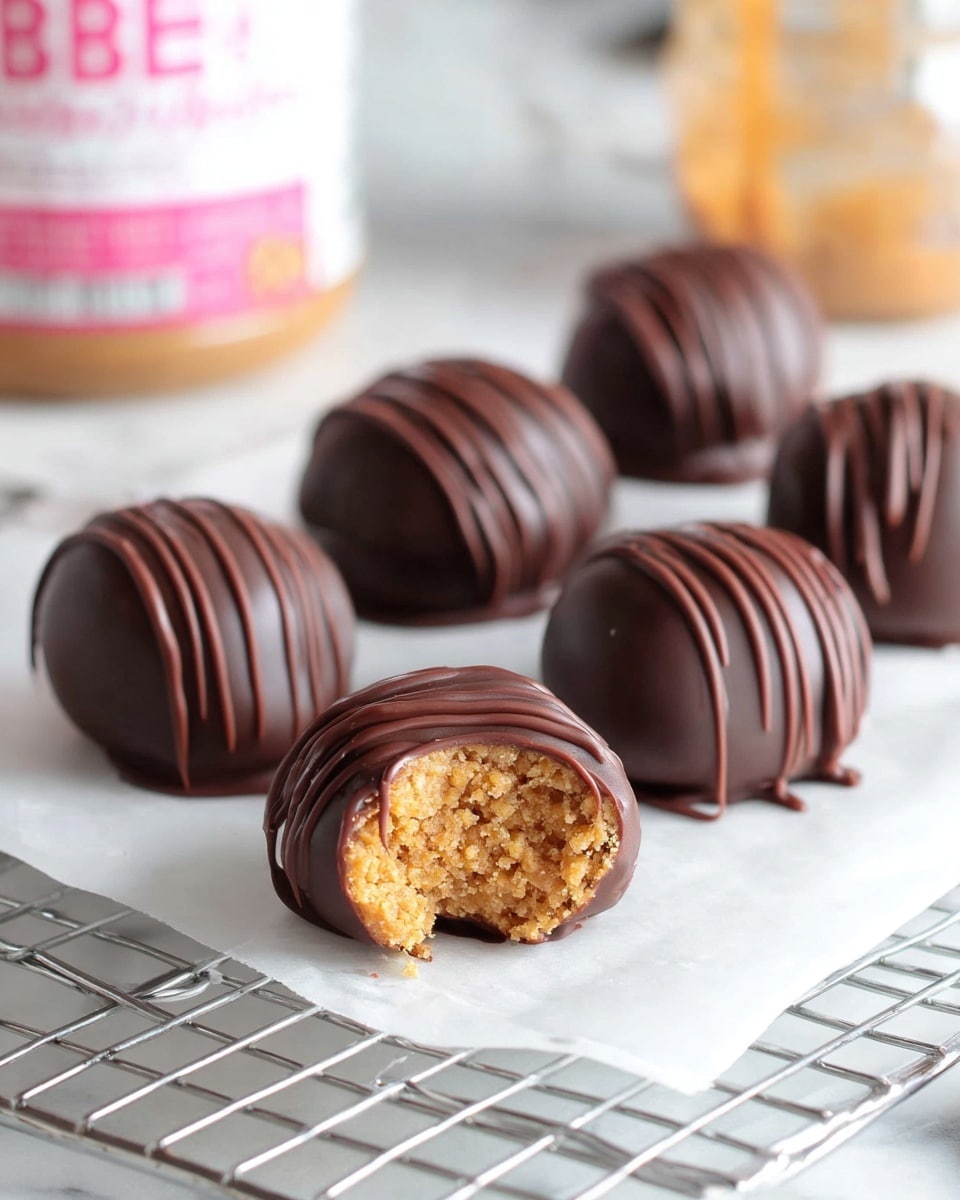 A close-up image of five chocolate-covered round treats resting on white parchment paper over a cooling rack, placed on a white marbled surface. Each treat is coated in smooth dark chocolate, with thin chocolate drizzle lines swirling on the top for decoration. One of the treats is bitten, revealing a golden brown, dense, crumbly filling inside. The background is softly blurred, showing a jar with a pink label and another container behind it. photo taken with an iphone --ar 4:5 --v 7