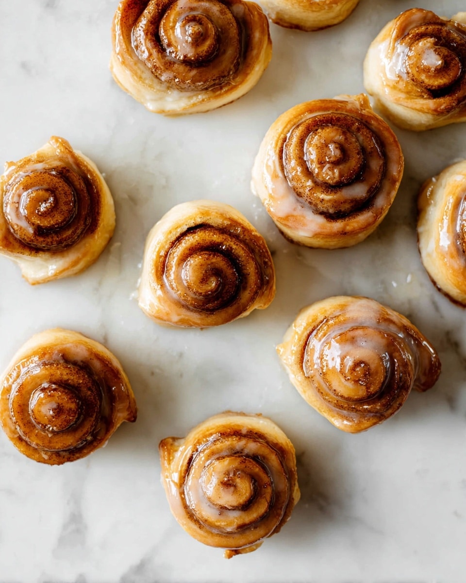 Several small cinnamon rolls are scattered on a white marbled surface. Each roll has a golden-brown outer layer with a slightly darker swirl of cinnamon inside, showing a spiral pattern on the top. The textured glaze is glossy and semi-transparent, pooling slightly around the base of many rolls, giving a sticky and shiny look. The rolls vary in height but all are round and compact. Photo taken with an iphone --ar 4:5 --v 7