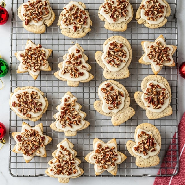 A variety of Christmas-themed cookies shaped like stars, Christmas trees, and mittens are laid out on a metal cooling rack placed on a white marbled surface. Each cookie has a light beige base speckled with tiny dark bits, topped with a creamy white icing layer that covers most of the top except the edges, with chopped nuts sprinkled on top adding texture and a mix of brown colors. The cookies have a slightly soft and smooth look from the icing, while the base appears firm and baked through. On the sides of the rack, there are small Christmas decorations in red and green, and a red and white cloth is partially visible on the white marbled surface. Photo taken with an iphone --ar 4:5 --v 7