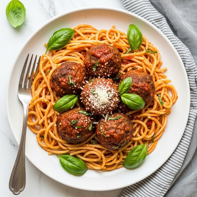 The dish shows a white plate filled with several layers of spaghetti noodles coated in red tomato sauce, with strands clearly twisted and mixed together at the base. On top of the noodles sit five large, round meatballs which have a browned texture and also some sauce on them. Bright green basil leaves are scattered around, adding spots of color, and a light sprinkling of grated white cheese can be seen on the center meatballs. A silver fork rests on the left side of the plate, which is placed on a white marbled surface with a soft gray striped cloth slightly visible on the side. Photo taken with an iphone --ar 4:5 --v 7