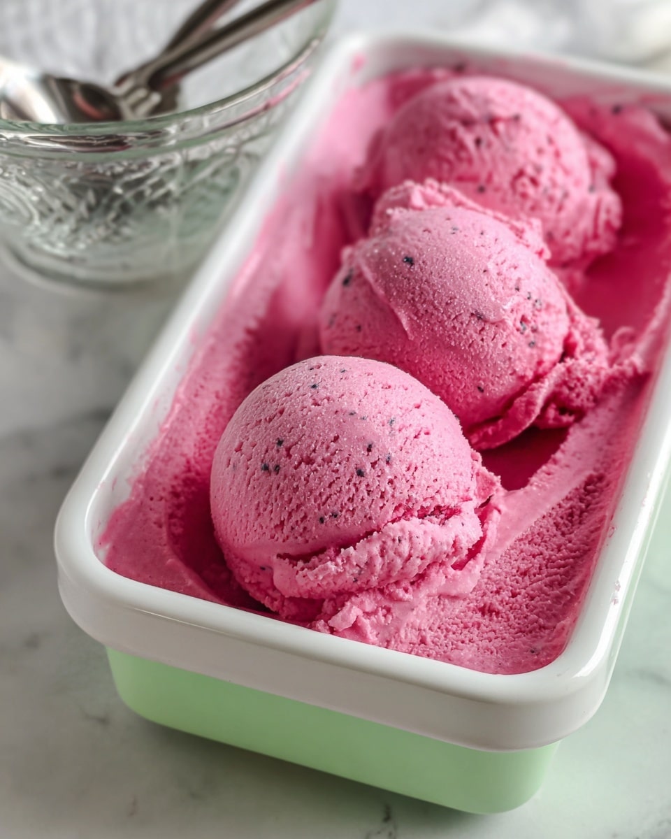 This image shows three scoops of bright pink ice cream with small dark specks in a white container with a light green lid. The ice cream is smooth and creamy with some texture from the small specks. The scoops sit on top of a bed of lightly disturbed ice cream of the same color and texture that fills the rest of the container. In the background, there is a glass bowl with two spoons, placed on a white marbled surface. Photo taken with an iphone --ar 4:5 --v 7