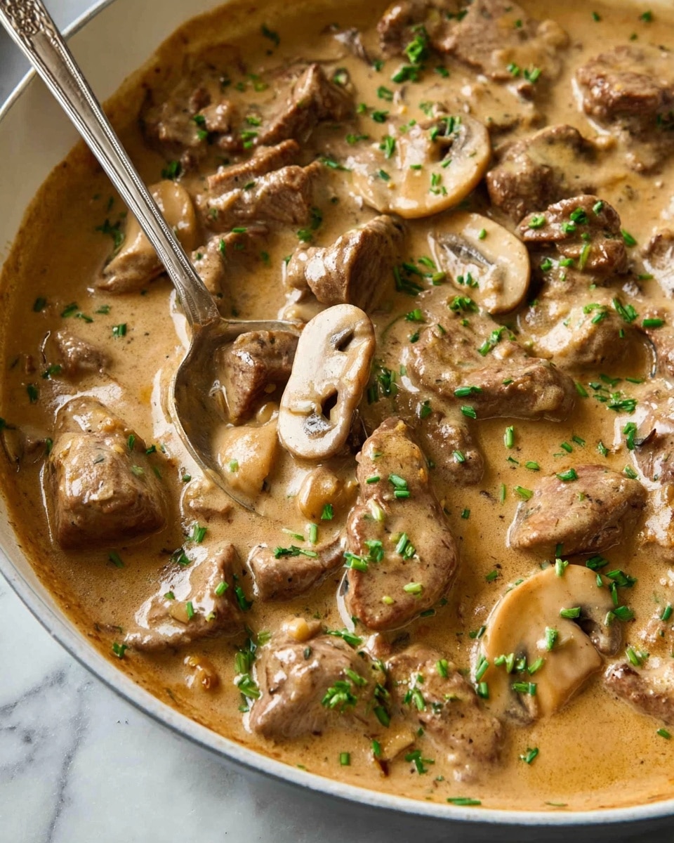 A close-up view of a creamy mushroom and beef dish in a white pan, showing a thick light brown sauce that covers tender pieces of sliced beef and mushroom slices. The beef pieces are spotted with light sear marks, and the mushrooms add a mix of smooth, pale beige textures. Small green chopped chives are sprinkled over the top, adding a touch of fresh color. A spoon rests inside the pan, partially submerged in the sauce. The pan sits on a white marbled surface. Photo taken with an iphone --ar 4:5 --v 7