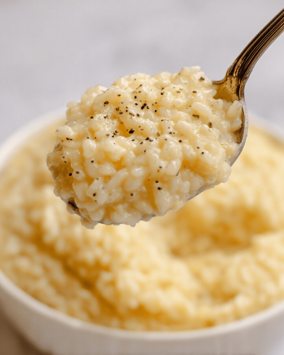 A close-up of a spoonful of creamy, pale yellow risotto with small black pepper flakes mixed throughout, held above a white bowl filled with more of the same risotto. The texture looks soft and slightly sticky, with the grains visible and clinging together. The background has a soft focus, showing the white marbled surface underneath the bowl photo taken with an iphone --ar 4:5 --v 7