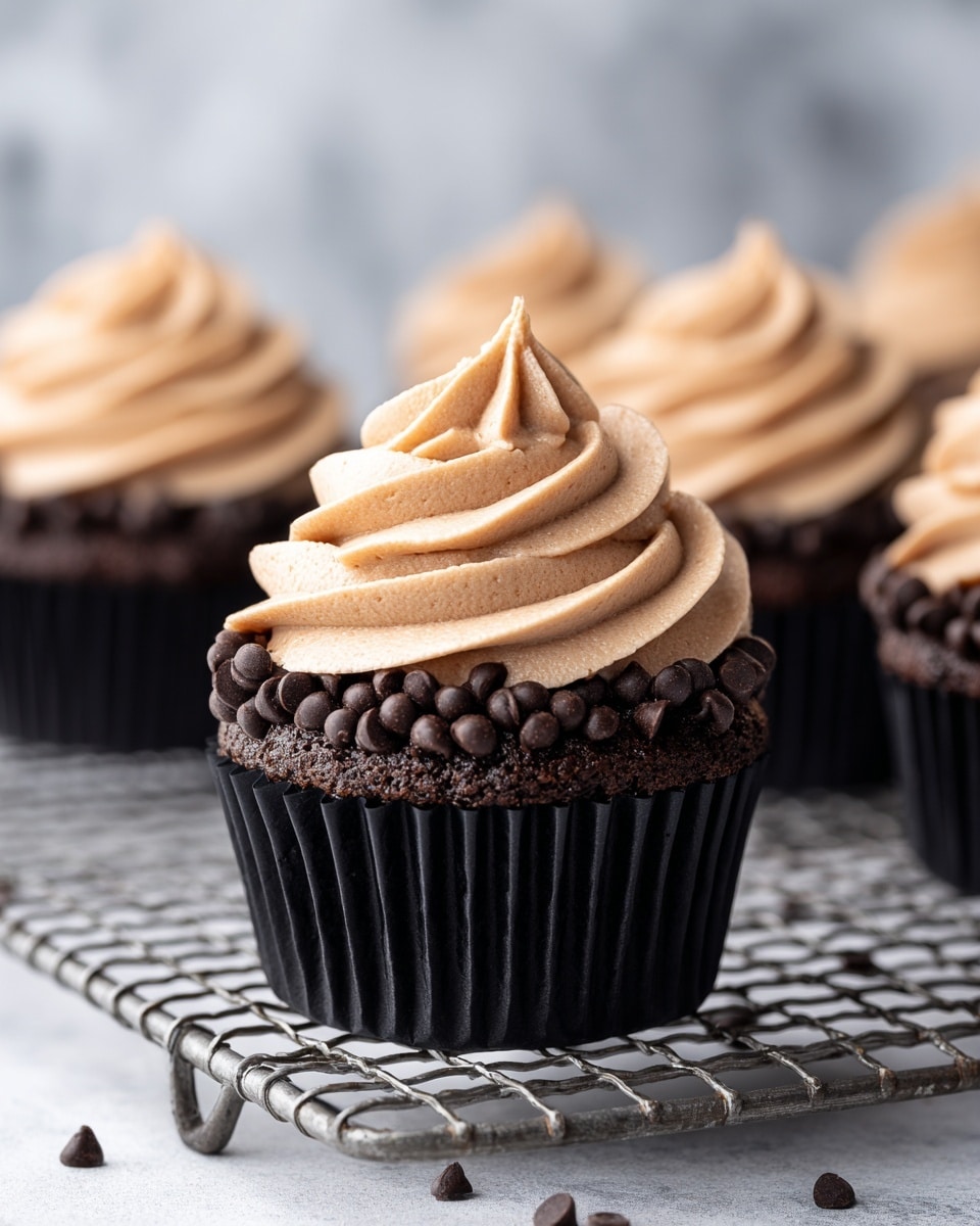 A dark chocolate cupcake in a black pleated wrapper sits on a metal cooling rack. The cupcake is topped with a thick, swirled layer of light brown frosting that spirals upward to a small peak in the center. Around the edge of the frosting, there is a ring of dark chocolate chips pressed into the surface, creating a textured border that contrasts with the smooth frosting. Additional cupcakes are blurred in the background on the same rack. The setting features a white marbled texture surface underneath. photo taken with an iphone --ar 4:5 --v 7