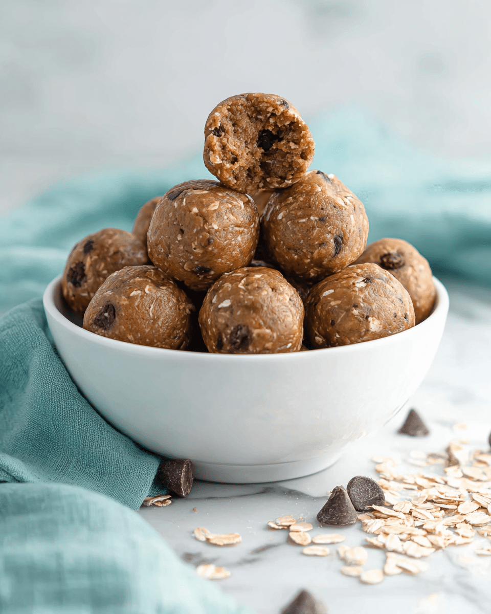 A white bowl filled with a pile of round, smooth-textured brown energy balls with visible small oats and dark chocolate chips inside. One energy ball is placed on top and tilted forward, showing its soft and chunky inside filled with oats and chocolate bits. The bowl rests on a white marbled surface with a soft teal cloth nearby, and a few scattered oats and chocolate chips lie in front of the bowl. photo taken with an iphone --ar 4:5 --v 7