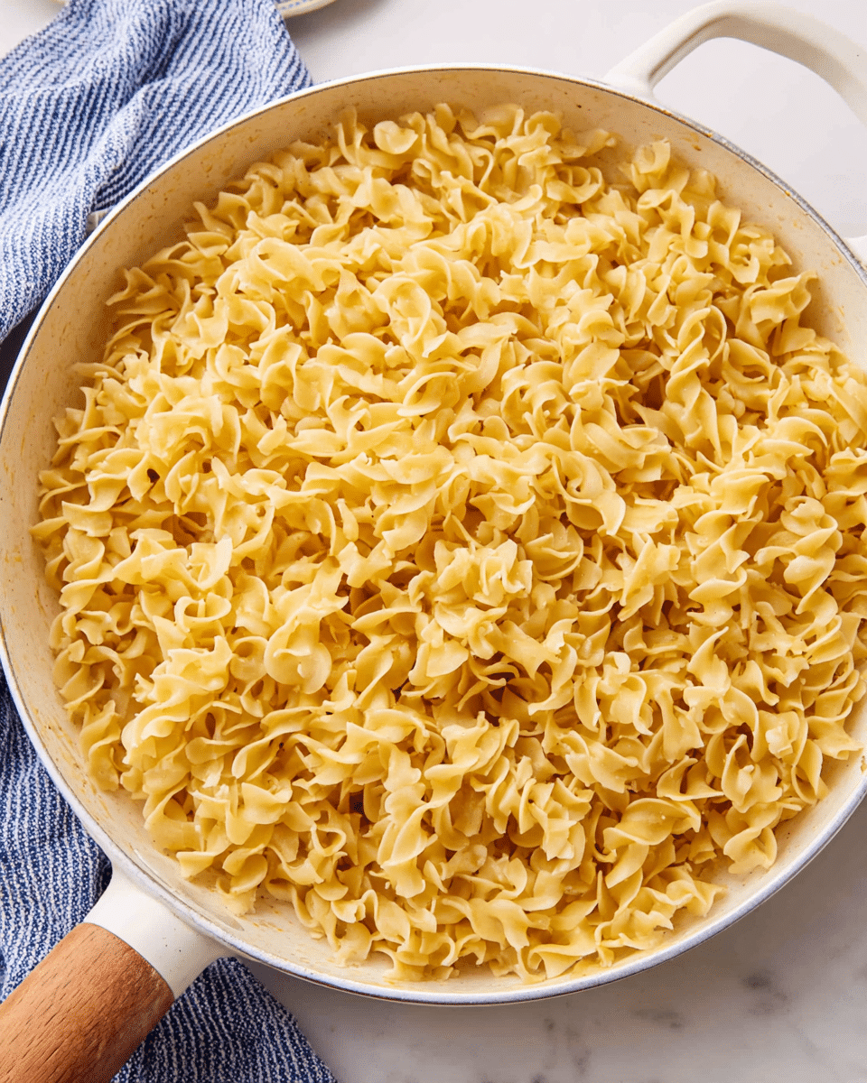 A close-up image showing a large white pan filled with one layer of cooked egg noodles. The noodles are golden yellow, curly, and have a soft texture with a slightly shiny surface. The pan has a white handle with a wooden grip, and it sits on a white marbled surface. A folded blue and white striped cloth is partially visible in the top left corner. Photo taken with an iphone --ar 4:5 --v 7