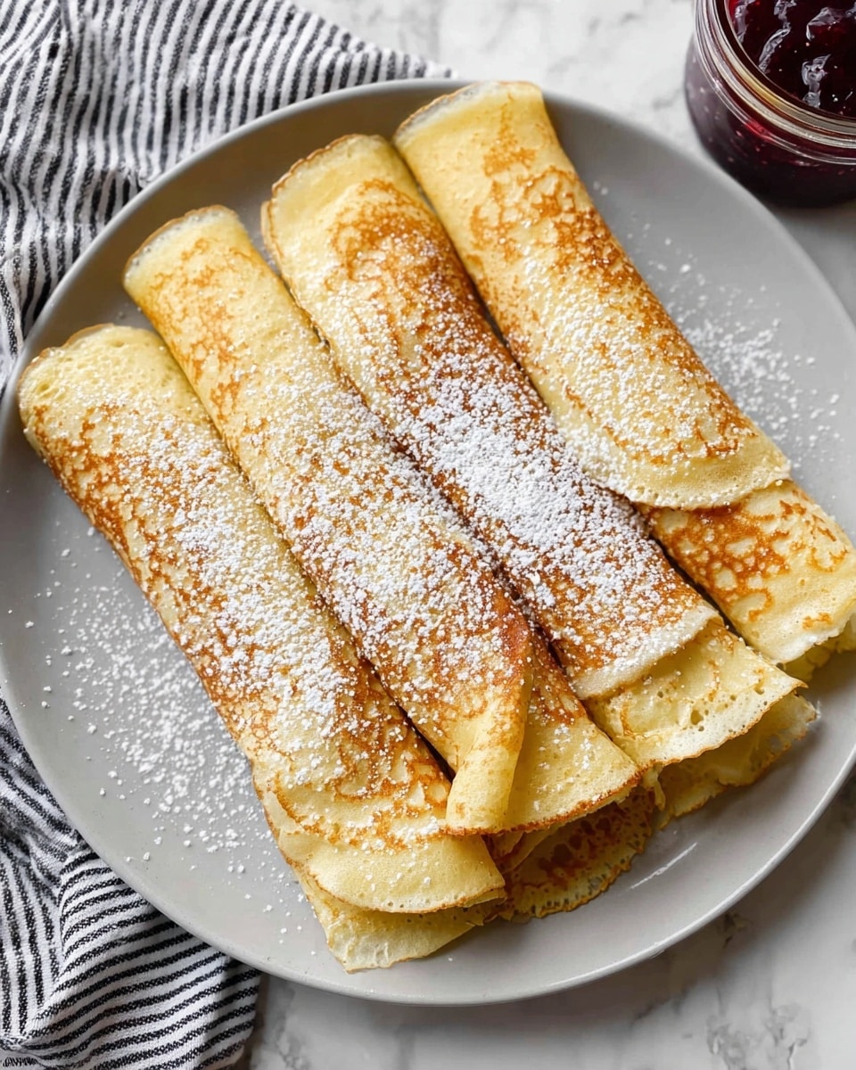 Five thin rolled crepes lie side by side on a round white plate, each crepe showing a golden-brown cooked surface with lighter yellow edges and dusted evenly with white powdered sugar. The crepes have a smooth texture with some small bubble marks and crispy spots where the batter cooked well. The round plate rests on a white marbled surface with a black and white striped cloth partially visible to the upper left. A small glass jar with dark red jam is placed near the top right edge of the frame. The overall look is simple and fresh, with the crepes as the clear focus. photo taken with an iphone --ar 4:5 --v 7