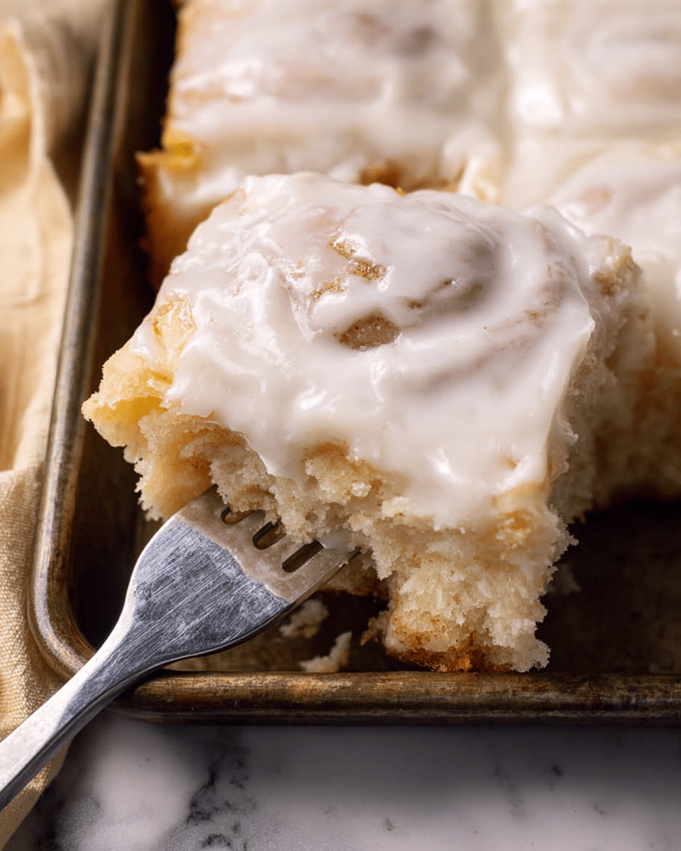 The image shows a piece of square cinnamon roll with white icing on top, held by a silver fork on a dark metal baking tray. The cinnamon roll is fluffy with a golden-brown color and visible swirls, covered in glossy, smooth white icing that has a slight shine. The background surface is a white marbled texture with a beige cloth partially visible on the left side. Photo taken with an iphone --ar 4:5 --v 7
