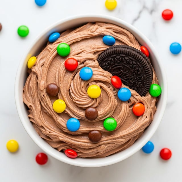 This dish shows a single-layer round dessert in a white bowl filled with thick, smooth swirl-textured milk chocolate mousse topped with scattered colorful candy-coated chocolates in red, yellow, green, blue, and brown. On one edge of the mousse, a half of a dark chocolate sandwich cookie is partially pressed into the creamy surface, adding contrast with its textured, embossed pattern. The bowl sits on a white marbled surface with more colorful candies scattered in the background, creating a lively and playful scene. photo taken with an iphone --ar 4:5 --v 7
