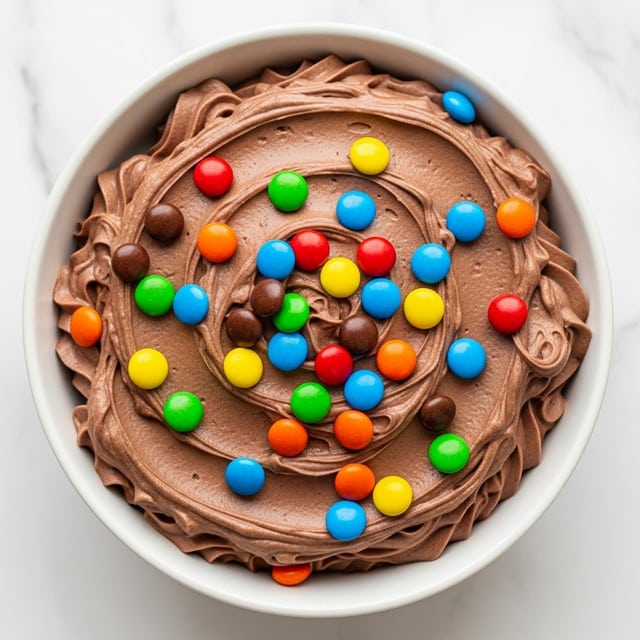 A thick layer of creamy chocolate mousse fills the white bowl, with soft swirls and peaks creating a smooth and rich texture. On top, colorful candy-coated chocolates in bright red, yellow, green, orange, blue, and brown are scattered, adding a vibrant contrast to the dark chocolate base. The white marbled surface under the bowl adds a clean, simple background that makes the dessert stand out clearly. photo taken with an iphone --ar 4:5 --v 7