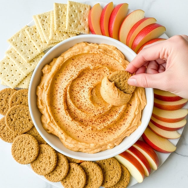 The image shows a white bowl filled with smooth, creamy, light orange dip sprinkled with a fine layer of red seasoning on top. Around the bowl, there are layers of beige square crackers, light brown round cookies, and pinkish-red apple slices with white inside arranged neatly on a white marbled surface. A woman's hand is dipping a light brown round cookie halfway into the dip. The overall scene is bright and clean, highlighting the textures and colors of the food. photo taken with an iphone --ar 4:5 --v 7