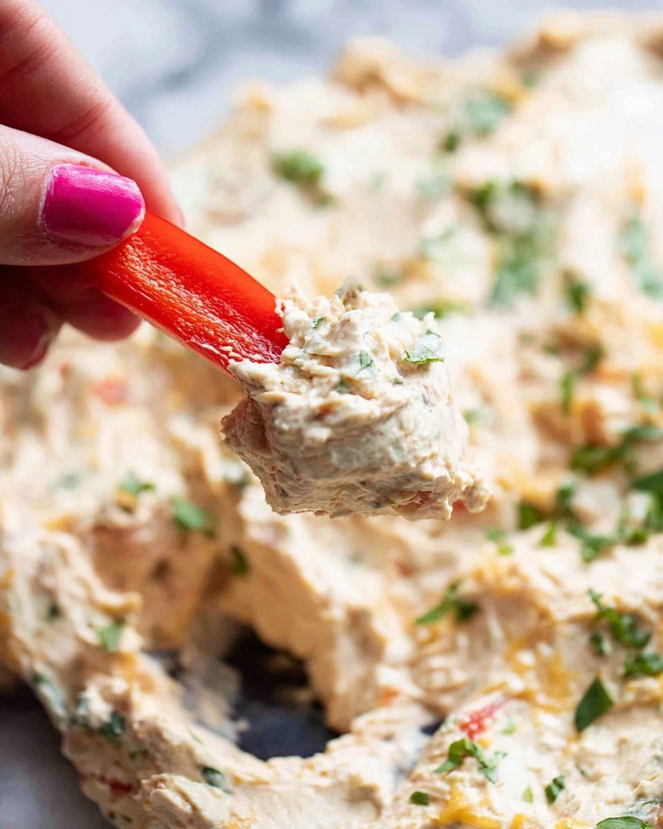 The image shows a close-up of a red bell pepper slice being held by a woman's hand with pink nail polish, dipping into a creamy, thick dip. The dip has a light beige color with green herb pieces sprinkled throughout, adding texture and color. The dip looks soft and spreadable, with a slightly chunky consistency visible in parts. The background is a white marbled surface with more of the creamy dip spread out, topped with small bits of green herbs and possibly melted cheese. Photo taken with an iphone --ar 4:5 --v 7