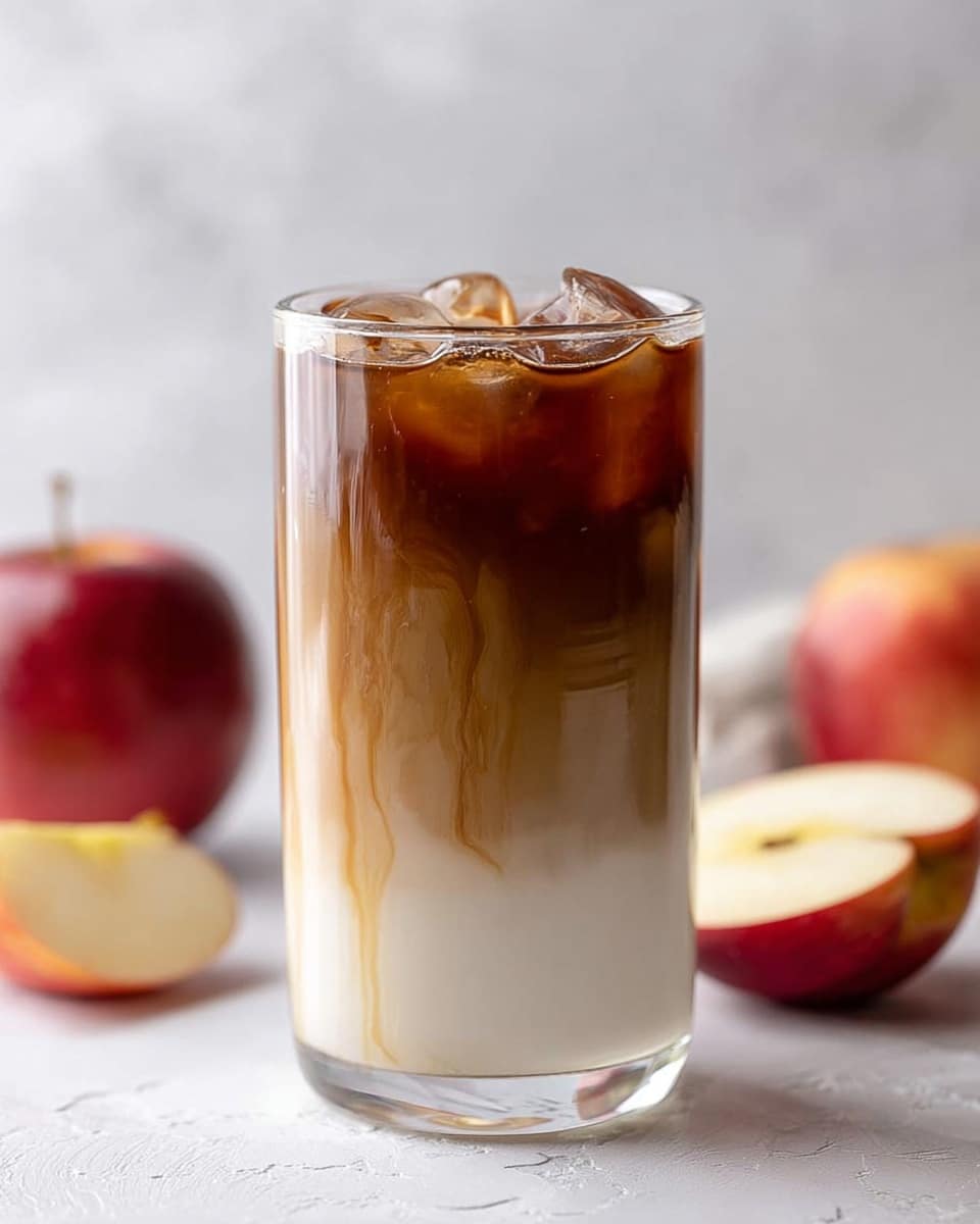The image shows a tall clear glass filled with a drink that has three visible layers. The bottom layer is a smooth white milk base. The middle layer is a light brown that looks like syrup slowly mixing with the milk, with streaks running down the sides of the glass. The top layer is dark brown iced coffee with ice cubes floating inside, giving it a refreshing look. The background is a soft white marbled texture. There are two red apples cut in half around the glass, one leaning on the left side and the other blurred in the background on the right. photo taken with an iphone --ar 4:5 --v 7