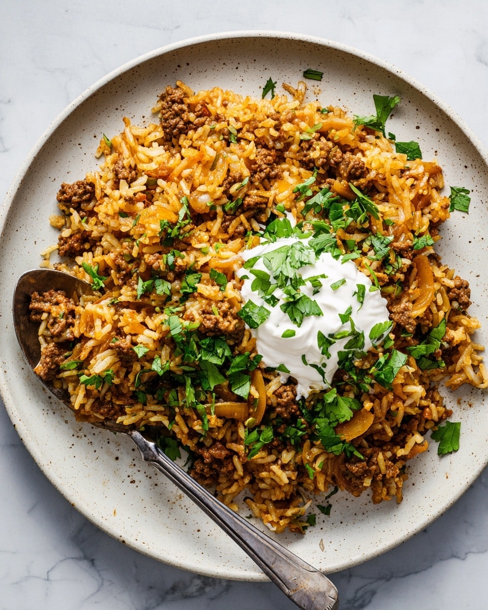 A close-up view of a single-layer dish served on a white plate with a subtle speckled texture, placed on a white marbled surface. The dish consists of cooked rice mixed with browned ground meat and sautéed onion slices, all in warm shades of brown and light orange. Scattered fresh green herbs, likely cilantro, add a lively contrast on top. In the center, there is a dollop of white sour cream garnished with more chopped green herbs. A silver spoon rests on the plate, ready to eat. Photo taken with an iphone --ar 4:5 --v 7