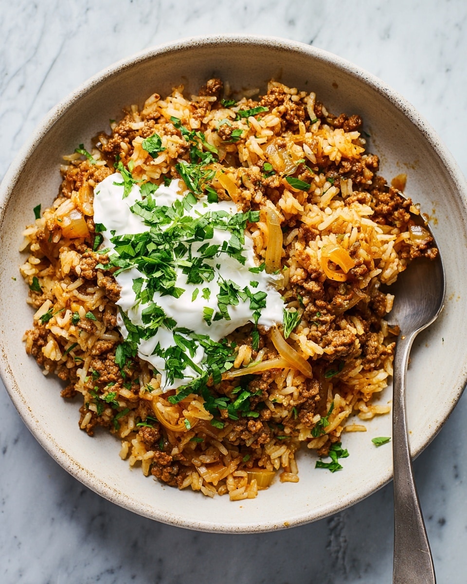 A bowl of cooked rice mixed with ground meat and sliced sautéed onions, showing a mix of light brown and orange colors. On top, there is a dollop of white sauce, likely sour cream or yogurt, sprinkled with fresh green chopped herbs. The dish is served in a white bowl with a rustic texture, placed on a white marbled surface, with a metal spoon partially inserted into the food from the bottom right side. Photo taken with an iphone --ar 4:5 --v 7