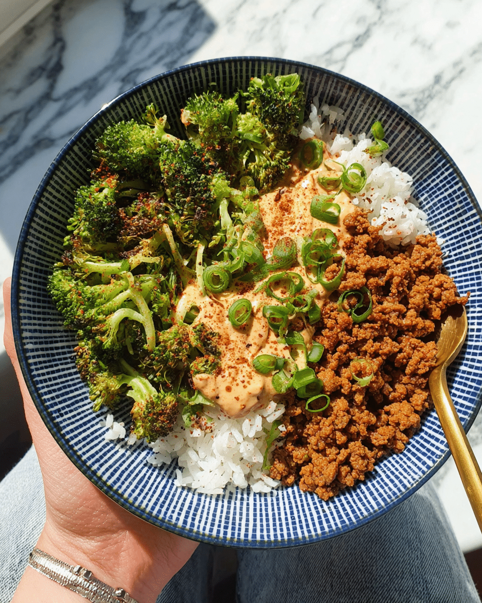 A bowl with a white base and blue grid pattern holds a colorful dish divided into three main parts: on the left, white fluffy rice topped with some crunchy fried bits and green sliced scallions; in the center, a dollop of light orange creamy sauce sprinkled with a bit of red spice and fresh green scallions; on the right, a heap of dark brown, textured cooked crumbles that look crispy; along the top edge, bright green roasted broccoli florets with a slightly charred look; a gold spoon is resting on the right side, and a woman's hand is holding the bowl against a white marbled surface, with soft natural light highlighting the food’s details. photo taken with an iphone --ar 4:5 --v 7