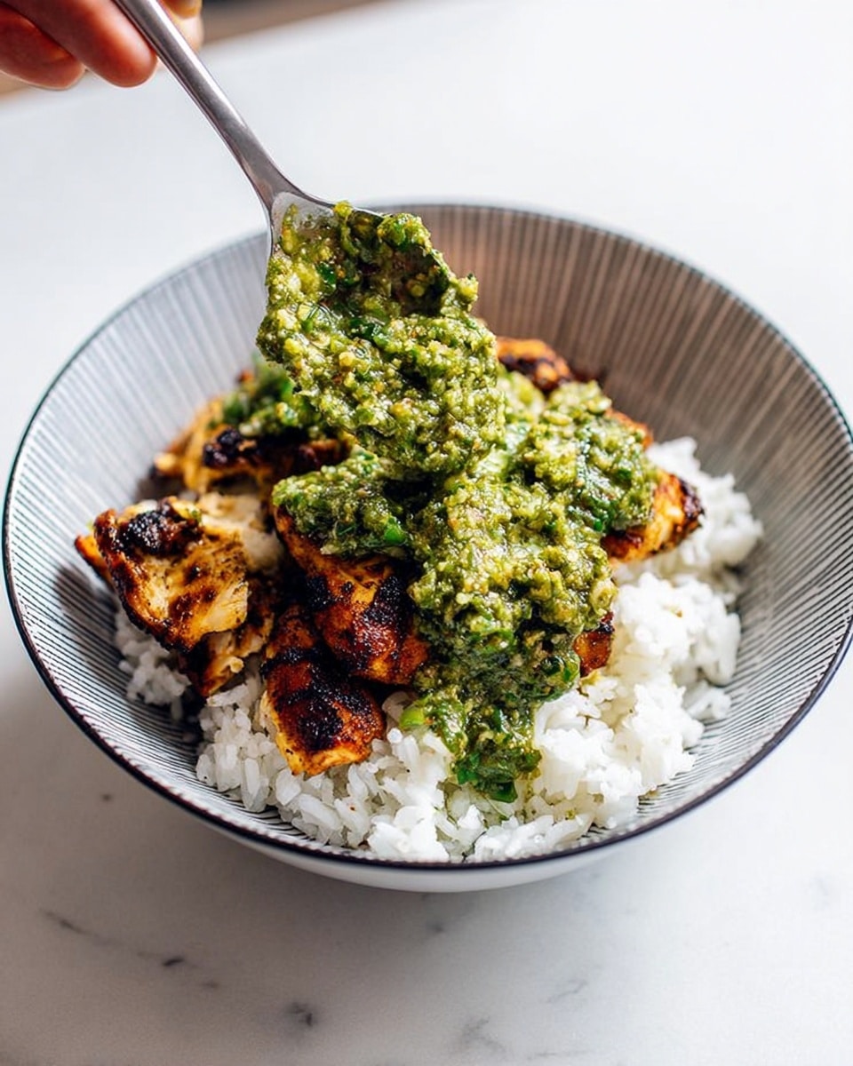 A bowl with white rice as the bottom layer, soft and fluffy with visible grains, topped with pieces of grilled chicken that are golden brown with char marks, covered unevenly by a chunky, green sauce that looks fresh and slightly textured. A silver spoon held by a woman's hand is adding the green sauce over the chicken. The bowl has a thin black rim and a subtle checkered pattern inside. The setting is on a white marbled surface. photo taken with an iphone --ar 4:5 --v 7