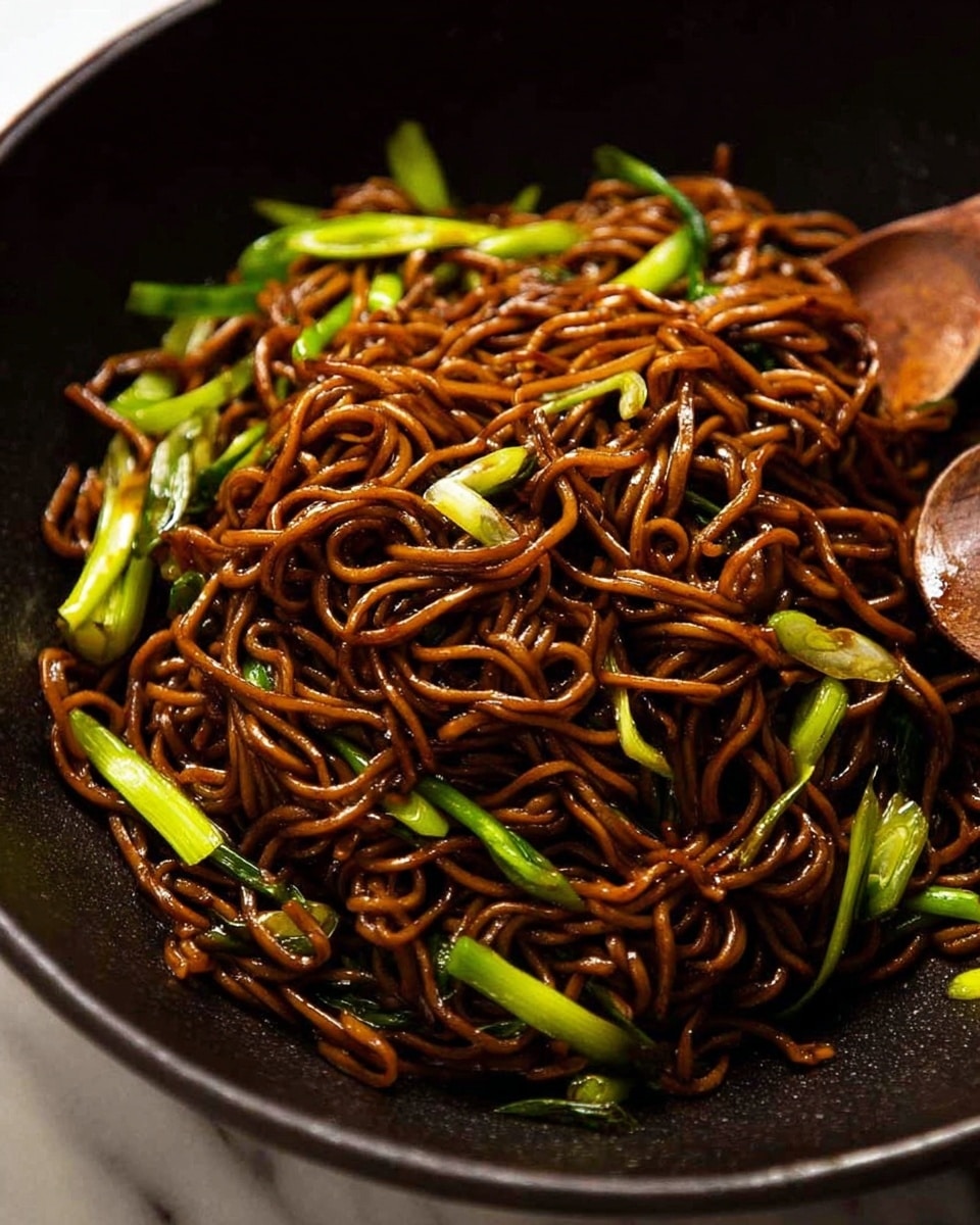 A close-up of a large black bowl filled with dark brown fried noodles mixed with bright green slices of vegetables, mostly scallions. The noodles look glossy and tangled with a slight shine, with the green vegetable pieces spread evenly throughout the dish, adding contrast. A wooden spoon is partially visible at the side, slightly covered by the noodles. The background is a white marbled surface. photo taken with an iphone --ar 4:5 --v 7