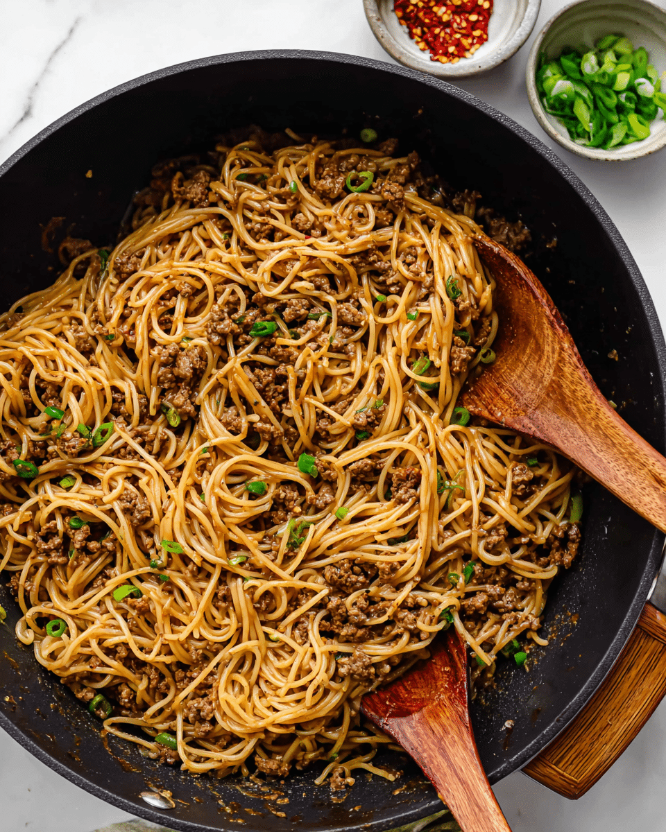 A large round black pan filled with cooked spaghetti noodles mixed with ground meat pieces and small green onion slices, showing a slightly oily texture with a light brown sauce coating the noodles and meat. Two wooden spoons are partially placed in the pan, partly hidden under the noodles, adding a warm natural wood tone. Small bowls with chopped green onions and red chili flakes sit at the upper edge of the frame on a white marbled surface, creating a clean, bright contrast to the dark pan. The scene is well-lit, focusing on the texture and color of the noodles and meat. photo taken with an iphone --ar 4:5 --v 7