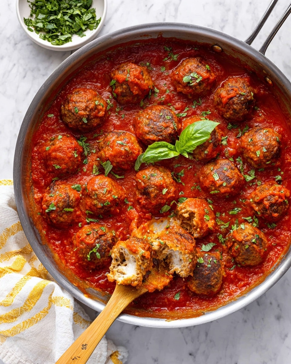 A large silver pan filled with round, golden-brown meatballs covered in thick, bright red tomato sauce, sprinkled with finely chopped green herbs. One meatball is cut open on a wooden spoon, showing a soft, melted white cheese center inside the meaty texture. A single fresh green basil leaf lies on the sauce beside the meatballs. The pan sits on a white marbled surface with a white bowl of chopped herbs and green basil leaves in the background, along with a white and yellow striped cloth nearby. Photo taken with an iphone --ar 4:5 --v 7