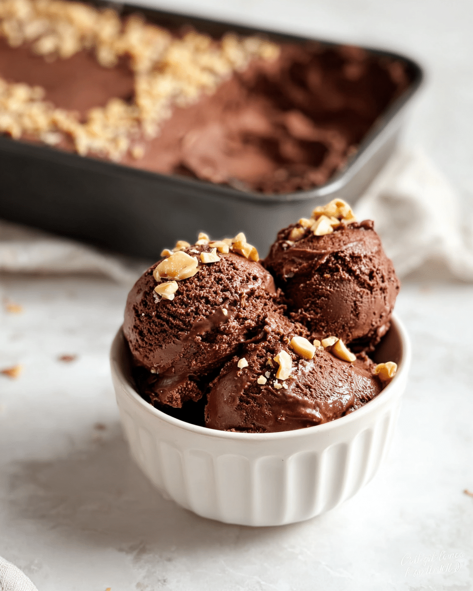 The image shows a white ceramic bowl filled with three scoops of rich, dark brown chocolate ice cream that has a smooth and creamy texture with small crunchy bits mixed in. The ice cream is topped with small pieces of light beige nuts scattered over the top. Behind the bowl, there is a pan filled with more chocolate ice cream topped with a thicker layer of nuts. The setting is on a white marbled surface. photo taken with an iphone --ar 4:5 --v 7