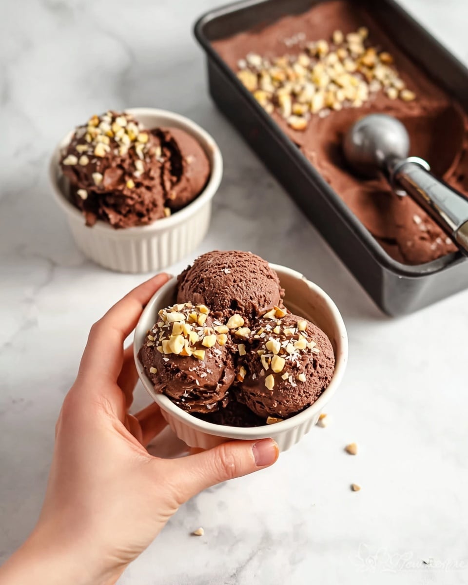 The image shows two small white ceramic bowls filled with dark brown chocolate ice cream, each bowl holding about three scoops with a rich, creamy texture. The ice cream is topped with a sprinkle of chopped nuts, adding a light beige color contrast on top. In the background, there is a black rectangular container partially filled with the same chocolate ice cream, also topped with chopped nuts, with a metal ice cream scoop resting inside. A woman's hand is holding one of the bowls in the foreground, set on a white marbled surface. Photo taken with an iphone --ar 4:5 --v 7