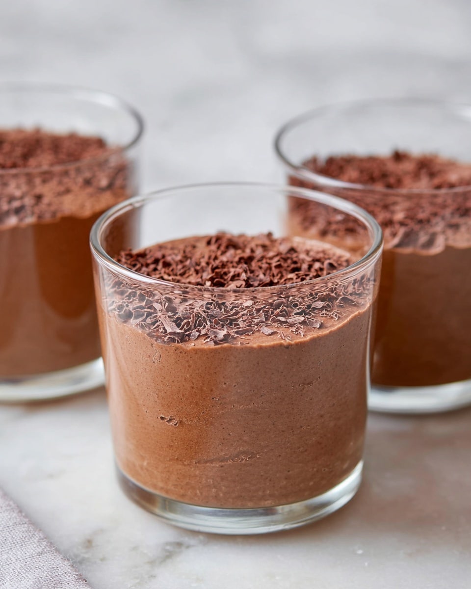 Three clear glass cups are filled with a rich chocolate mousse, each with one smooth, thick layer of light brown mousse topped with finely grated chocolate shavings. The mousse looks soft and creamy, filling the glasses almost to the top. The cups are arranged in a row on a white marbled surface, with a soft, blurred background. The middle and back cups are slightly out of focus, drawing attention to the closest one. photo taken with an iphone --ar 4:5 --v 7